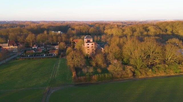 golden hour bornem centers tall brick tower dense woodland beside green fields warm sunlight treetops nearby village homes along forest&rsquo;s edge from rising drone view belgium forest 
