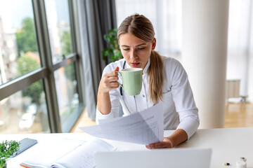 Female doctor in white coat reading medical documents and drinking coffee at desk, healthcare and medicine concept. Professional physician reviewing patient reports and test results in home office. © Graphicroyalty