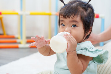Adorable asian toddler girl drinking milk from bottle while sitting on soft mat at home during playtime moment of early learning and family bonding showing curiosity growth and comfort indoors