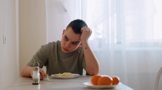 Unhappy boy sits at dining table with plate of food untouched, resting head on hand with sad expression, natural daylight through curtain capturing moment of refusal and frustration