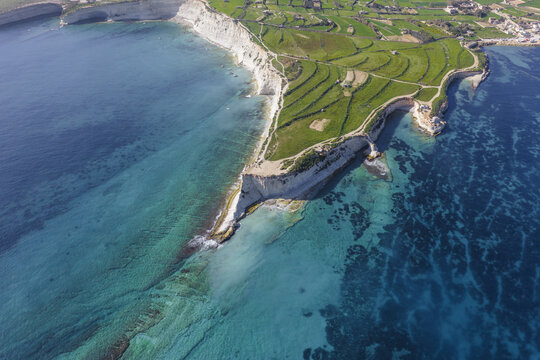 Aerial view of Munxar Path cliffs contrast with the deep blue sea near the green fields of Marsaskala, Marsaskala, Malta.