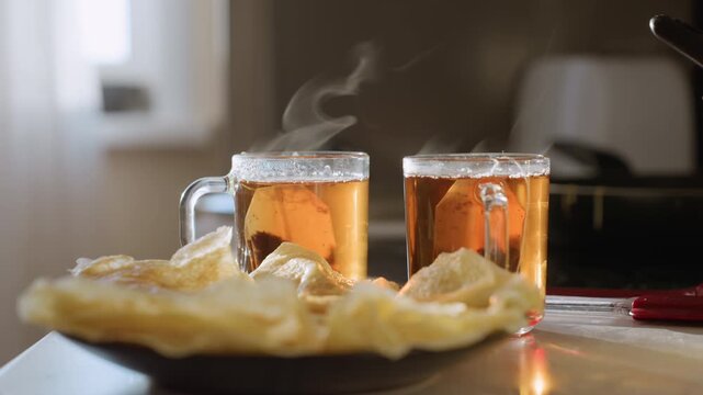 Close up of two steaming cups of herbal tea with teabags inside, vapor rising above hot surface, pancakes served on plate in foreground, warm morning breakfast setting