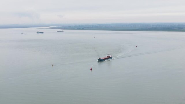 small coastal cargo vessel or inland freighter travels across dutch river channel weaving between red yellow navigation buoys leaving narrow curved wake calm water wide netherlands 