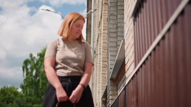 Tired lady walking toward brick building under warm summer sunlight, holding bag and stopping to rest against wall, expressing exhaustion, quiet thought, and emotional pause