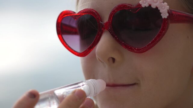 Girl lip gloss application. Young child applying clear shiny lip balm outdoors while wearing bright red heart sunglasses.