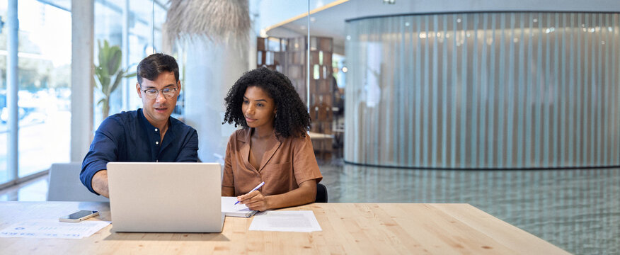 Busy team professional employees working together on computer discussing online technology data at work. Two young diverse coworkers business people using laptop and talking at office meeting.