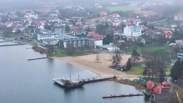 misty perspective charzykowy poland lakeside pier moored boat quiet sandy beach fog softened town buildings muted greenery revealing aerial drone air mist water shoreline dock harbor 