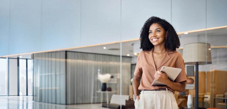 Happy confident young African American professional woman, female entrepreneur, businesswoman looking away holding tab standing in office at work dreaming of future success.