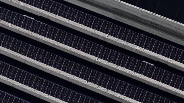 Diagonal rows of photovoltaic solar panels cover a large industrial buiilding rooftop, creating a repeating geometric pattern of dark modules and metal supports for renewable electricity generation