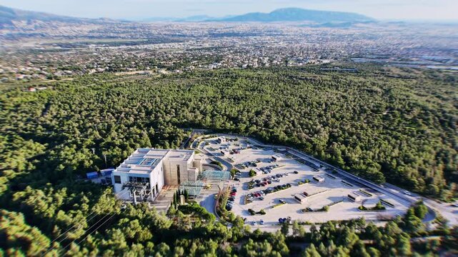 100 meters altitude from near Regency Casino Mont Parnes, the drone captures the mountain base with prominent parking area and cable car building below. Dense lush forest envelops the surroundings