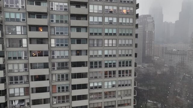 Aerial view of downtown American metropolis during winter snowstorm, residential high rise tower with lit windows, foggy skyline in background, cold urban atmosphere. Orbit shot.