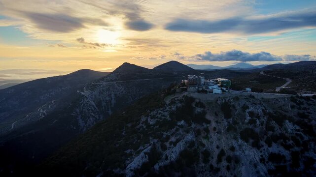1100 meters altitude, the drone moves rightward capturing Regency Casino Mont Parnes atop its mountain peak in the frame's distance. Jagged mountaintops rise ahead bathed in golden sun.