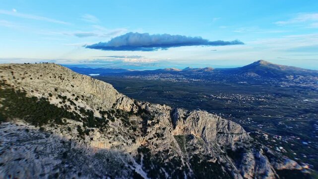 Drone glides leftward from the mountain side hosting Regency Casino Mont Parnes, capturing the rocky summit in frame. Beyond stretches a vast plain leading to distant sea under blue sky