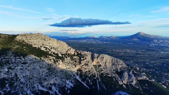 Starting from above the Mont Parnes summit, the drone moves capturing the jagged rocky peak prominently in frame. A sweeping plain unfolds behind leading toward the far-off sea horizon