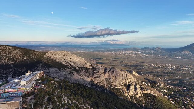 Hovering above Regency Casino Mont Parnes on Mount Parnitha, the drone pulls back slowly with the casino positioned to the left. A rugged rocky mountain looms directly ahead, perfectly framed by a sol