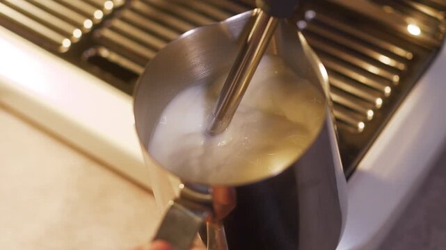 Slow motion close up of milk being frothed in a stainless steel pitcher using a steam wand on a portafilter espresso machine. Warm lighting highlights swirling foam texture and barista preparation pro