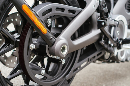 A high-detail shot of the rear powertrain of an electric motorcycle. The image showcases the carbon drive belt, sprocket, and rear swingarm in a matte grey finish, along with an orange safety reflecto