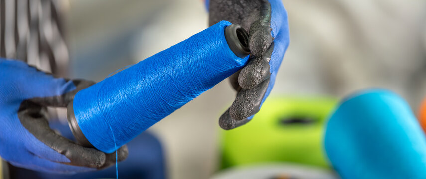 Close up of worker hands in gloves holding blue yarn spool with colorful thread cones background in textile factory. Collection of multicolored sewing materials for weaving and knitting.