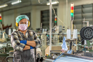 Fototapeta premium Confident textile factory worker posing with arms crossed in weaving machine mill. Portrait of male industrial staff wearing face mask and ear protection standing in fabric manufacturing plant.