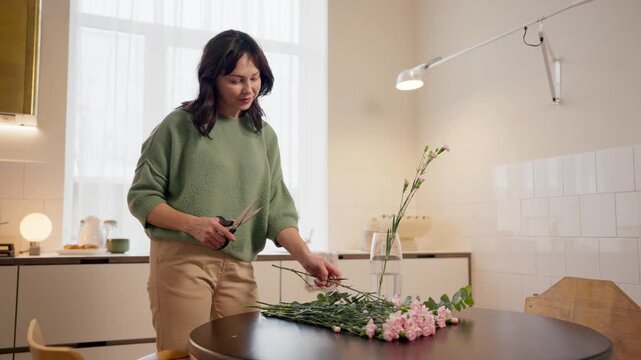 Caucasian mother standing in kitchen. Adult female cutting flower stems with scissors. Arranging bouquet into glass vase. Preparing gift for family. Focused woman enjoying moment at home. Interior.