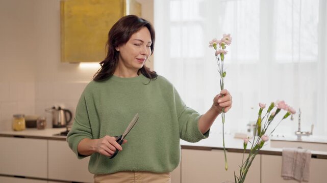 Charming mother standing in kitchen. Woman cutting flower stems with scissors. Placing flowers into glass vase. Smiling while preparing home decor. Family care concept at home. Daytime interior scene.