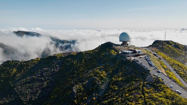 Aerial view of Pico do Areeiro weather radar station rising above clouds, mountain road and rugged terrain visible in high altitude landscape, Madeira Island, Portugal.