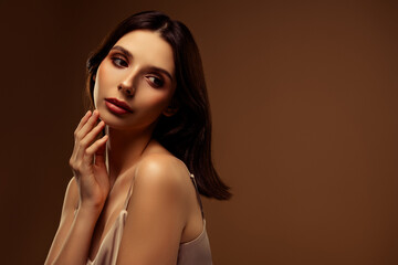 Attractive young woman with brunette hair gracefully posing against a beige background wearing satin
