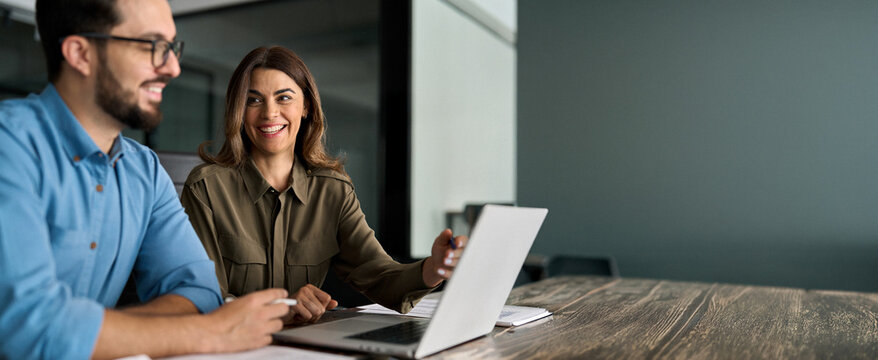 Happy busy female manager explaining corporate software work talking to young male employee consulting client working on computer using laptop discussing business project plan at office meeting.