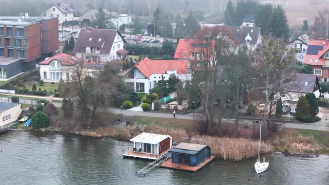 fog muted lakeside charzykowy poland red roofed village homes floating modern cabins lone white sailboat gently rippled water under calm aerial sky drone air waterfront residential roofs 