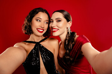 Two glamorous women in elegant red and black dresses pose for a selfie at a festive party