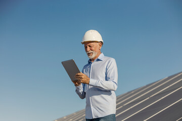 Senior Environmentalist Wearing Hard Hat Inspecting Solar Panels Using Tablet Outdoors