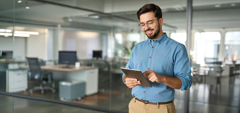 Smiling busy young latin business man manager using tablet computer, happy hispanic businessman executive looking at tab device analyzing finance trading market data working standing in office.