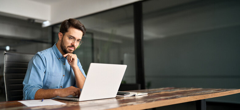Busy professional business man company employee, young male programmer, latin businessman looking at computer thinking on online investment or software development using laptop working at office.