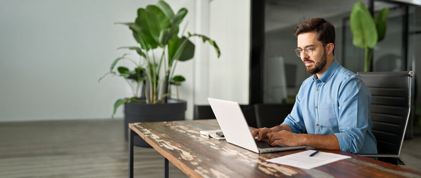 Busy professional business man executive, young male corporate worker, latin businessman wearing glasses typing on computer technology using laptop searching on web working at office workplace desk.