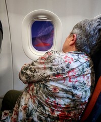 Elderly woman traveling on an airplane sitting by the window. Adult woman sitting in the front window of an airplane. Female traveler looking out the window. Elderly tourist traveling on an airplane.