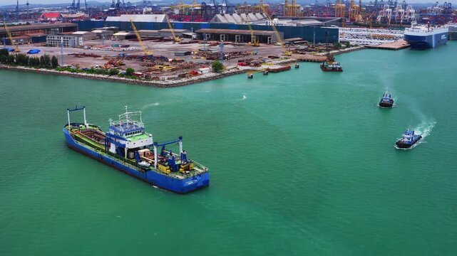 A large blue container ship filled with multicolored cargo containers sails through the ocean, followed by a small escort vessel.