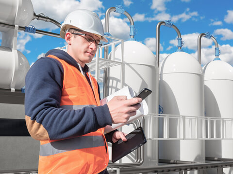 LNG facility inspection. Engineer in safety gear uses smartphone and tablet to check storage tanks and piping systems, monitoring operations, maintenance and safety at liquefied natural gas plant.