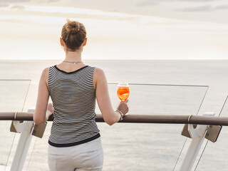Stylish woman with a glass of drink standing on the empty deck of a cruise ship against the backdrop of sea waves. Sunny day. Chill lifestyle. View from the back. Perfect for holiday, travel themes © Svetlana