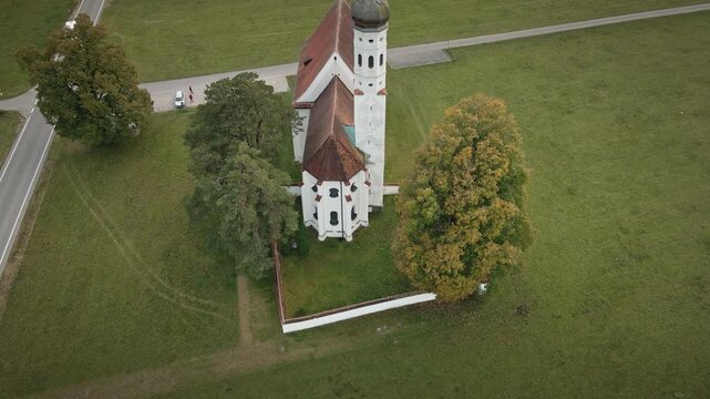Smooth 4K drone descends by St. Coloman Church in Schwangau as camera tilts up, shifting focus from roofline to tower and open landscape beyond.