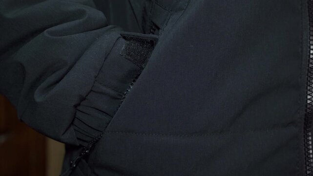 A man put his hand into the side pocket of a black jacket,  A man warms his hands in the pockets of his jacket, A hand in the pocket of a black tactical jacket with velcro cuffs