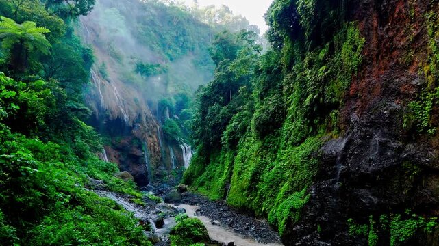 Drone flies through Tumpak Sewu canyon in Java, Indonesia, revealing lush green walls, mist, small waterfalls, winding riverbed, and serene tropical landscapes in 4K.