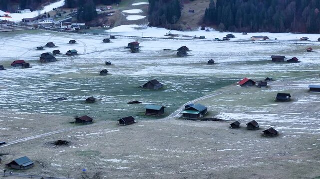 alpine meadow garmisch partenkirchen dotted small wooden hay barns scattered across patchy snow winter grass connected by faint farm tracks beneath forested mountain near garmisch-partenkirchen 