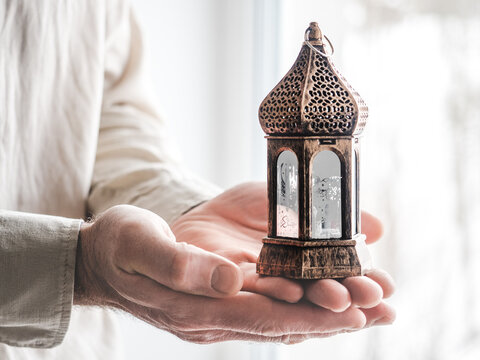 Man's hands hold a traditional Ramadan lantern with gold calligraphy, mosque motifs and star patterns. Soft glow, symbolic of faith, peace and Eid celebration