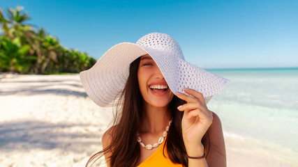 Portrait of young woman wearing straw hat standing at beach with light blue ocean in background, lady smiling at camera, walking on sandy beach © Home-stock