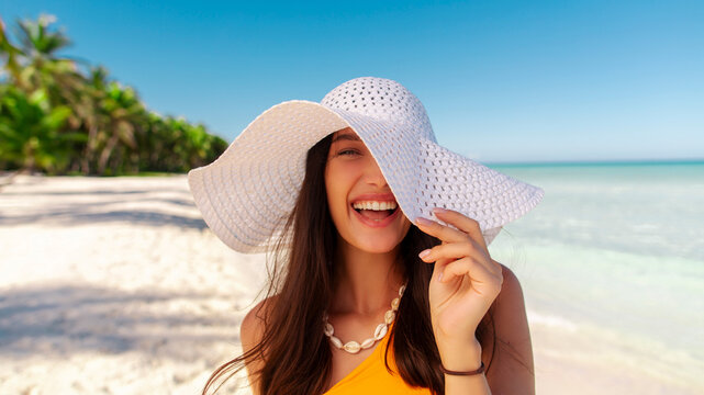 Portrait of young woman wearing straw hat standing at beach with light blue ocean in background, lady smiling at camera, walking on sandy beach