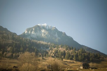 A stunning mountain rises high, its peaks dusted with snow, surrounded by lush green forests and rolling hills. The calm valley below glistens in the warmth of the sun