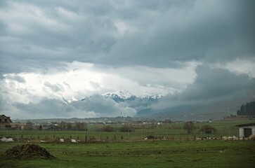 Obraz premium Clouds swirl over the breathtaking mountain range while a peaceful countryside unfolds below. Farms and fields blend harmoniously with nature's artistry, creating a serene scene