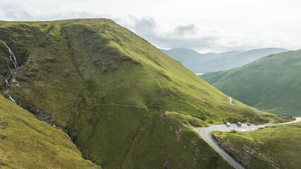 Fototapeta premium Scenic mountain pass with winding road and parked vehicles, showcasing lush green slopes and distant hazy peaks