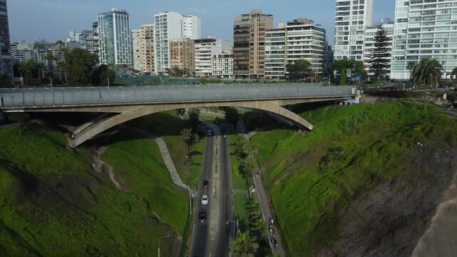 Aerial shot of a bridge over a road called "Bajada Balta". Done flies backwards and up shoting the coastal road named "Costa Verde" and the many buildings of the Miraflores district of Lima, Peru.