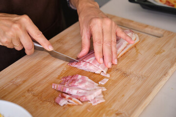 Hands slicing raw bacon into small pieces on a wooden cutting board in a kitchen